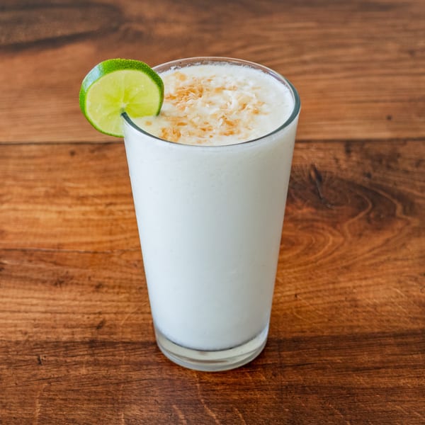 A tall glass of white creamy drink, garnished with a lime slice and toasted coconut flakes.