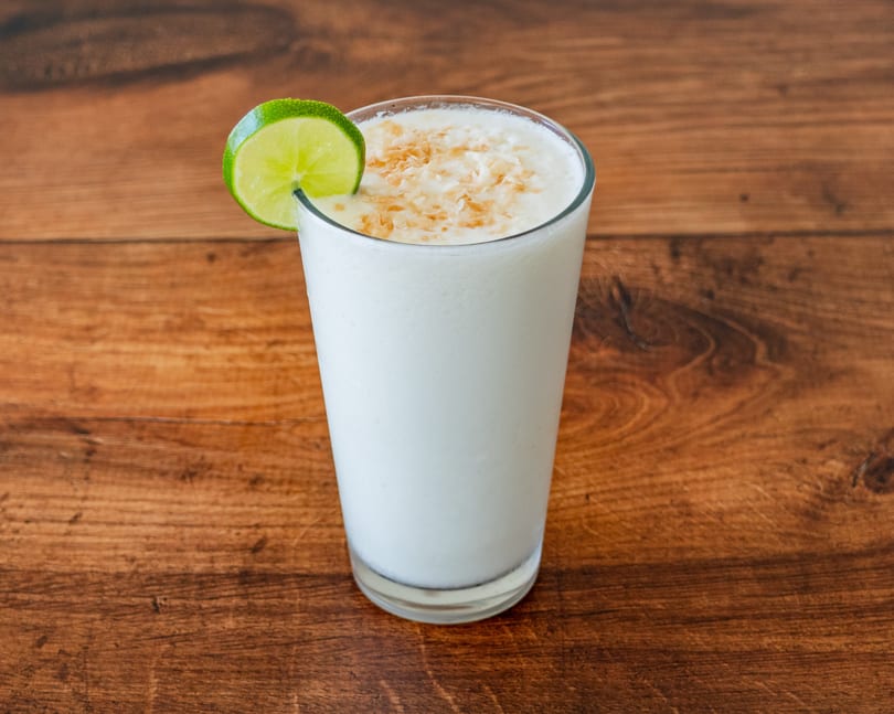 A tall glass of white creamy drink, garnished with a lime slice and toasted coconut flakes.