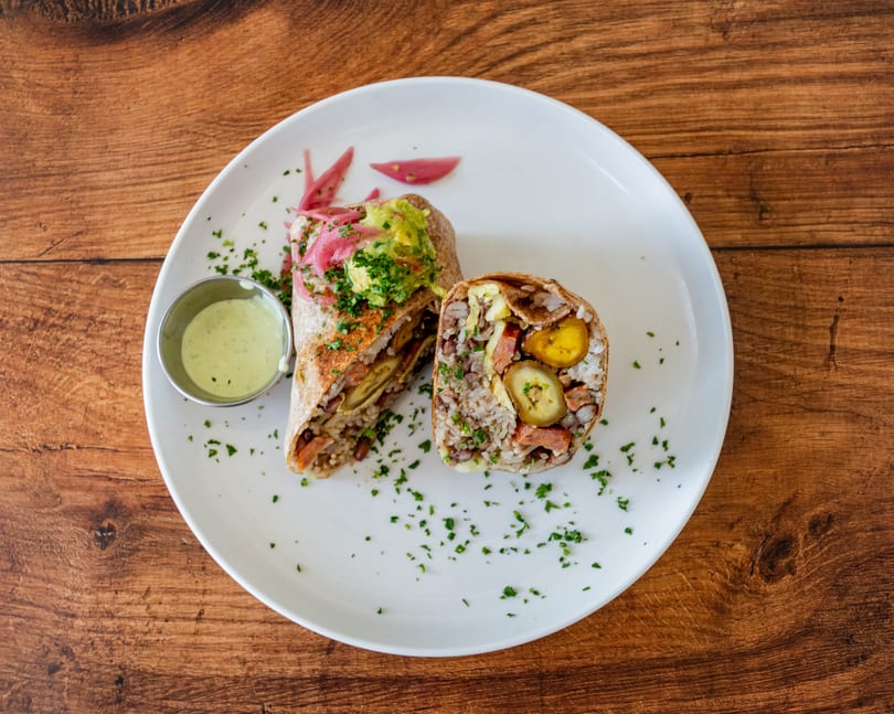 A sliced burrito with rice, beans, plantains, and sausage, topped with guacamole and pickled onions.