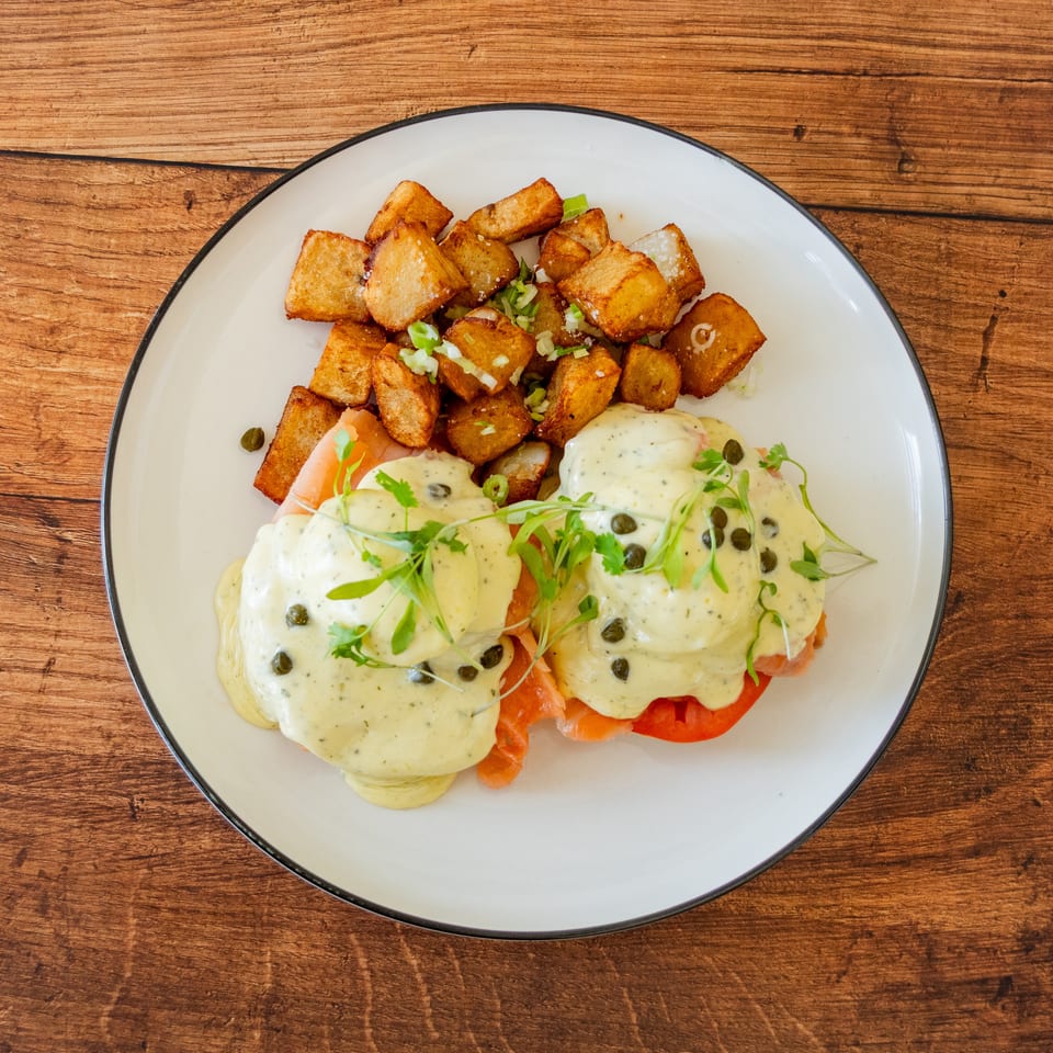 Smoked salmon eggs Benedict with capers and microgreens, served with crispy home fries on a white plate.