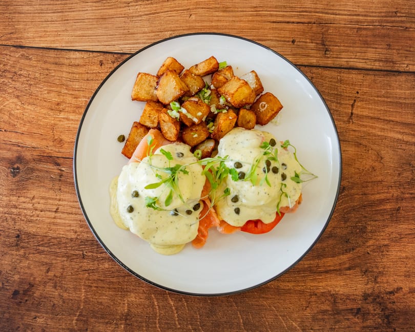 Smoked salmon eggs Benedict with capers and microgreens, served with crispy home fries on a white plate.
