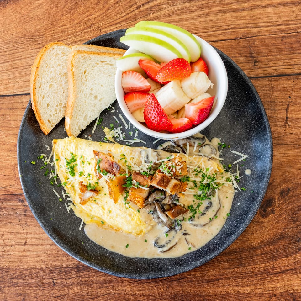Omelet with mushroom sauce, bacon, cheese, parsley, toast, and a fruit cup on a wooden table.
