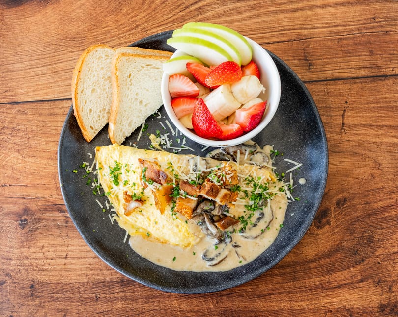 Omelet with mushroom sauce, bacon, cheese, parsley, toast, and a fruit cup on a wooden table.