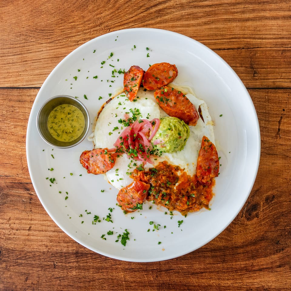 Breakfast plate with fried eggs, chorizo, guacamole, salsa, pickled onions, and a side of green sauce.