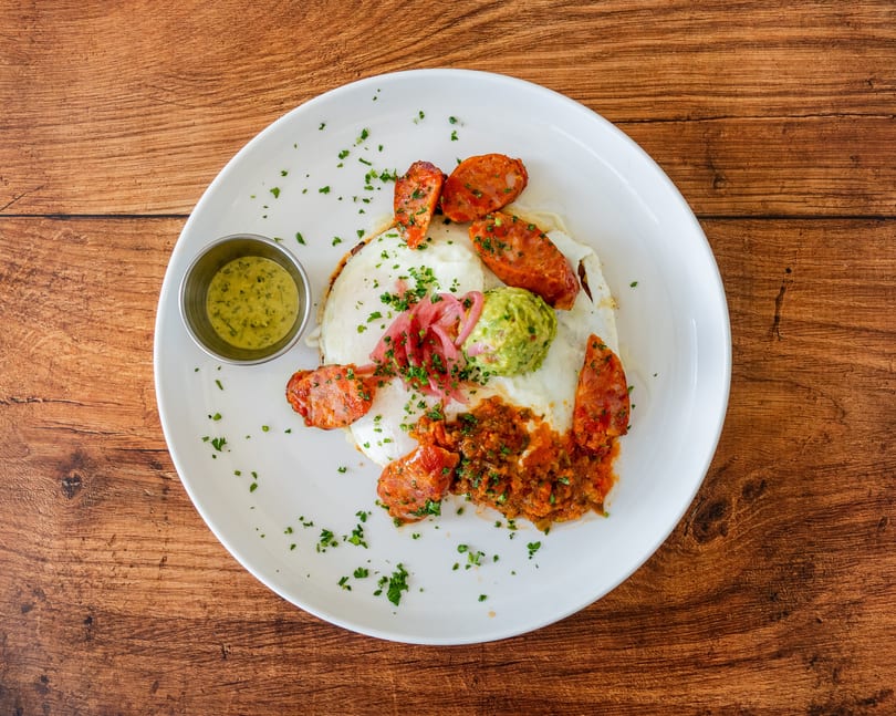 Breakfast plate with fried eggs, chorizo, guacamole, salsa, pickled onions, and a side of green sauce.