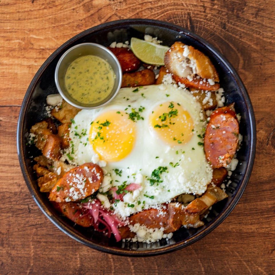 Two fried eggs with chorizo, potatoes, cheese, and a side of green sauce on a wooden table.