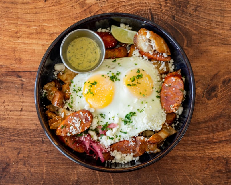 Two fried eggs with chorizo, potatoes, cheese, and a side of green sauce on a wooden table.