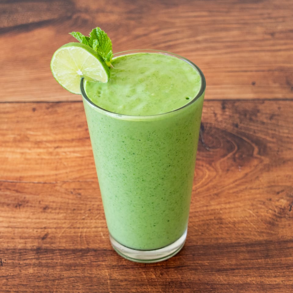 Green smoothie in a tall glass, garnished with a lime slice and mint, on a wooden table.
