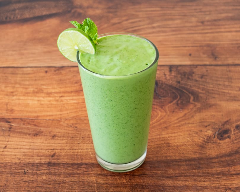 Green smoothie in a tall glass, garnished with a lime slice and mint, on a wooden table.