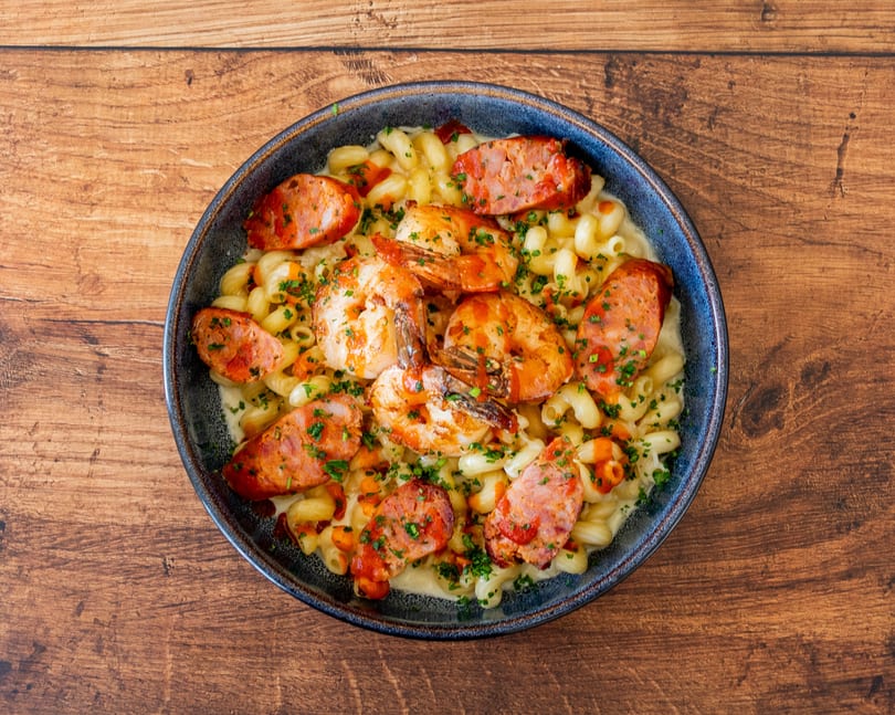 Macaroni and cheese with shrimp and sliced sausage, garnished with parsley, on a wooden table.