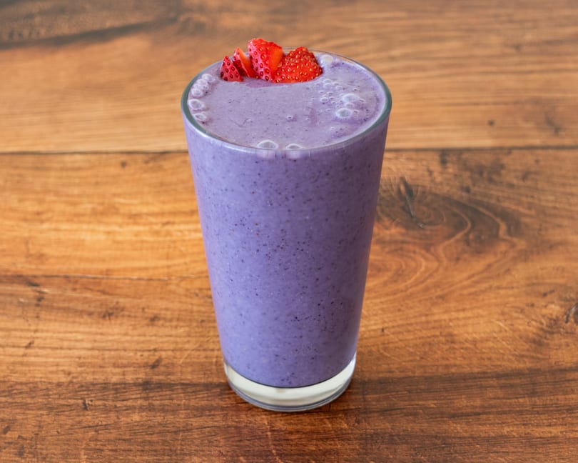 A tall glass of purple berry smoothie, topped with sliced strawberries, on a wooden table.