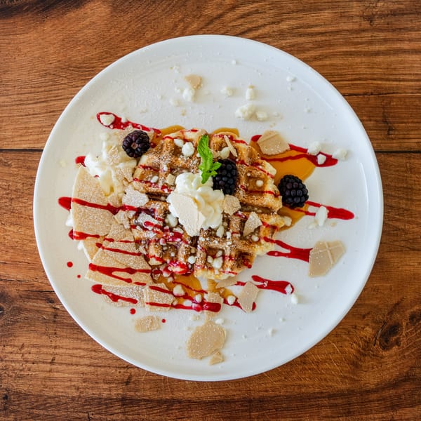 A waffle dessert with whipped cream, blackberries, mint, and berry and caramel drizzles on a white plate.