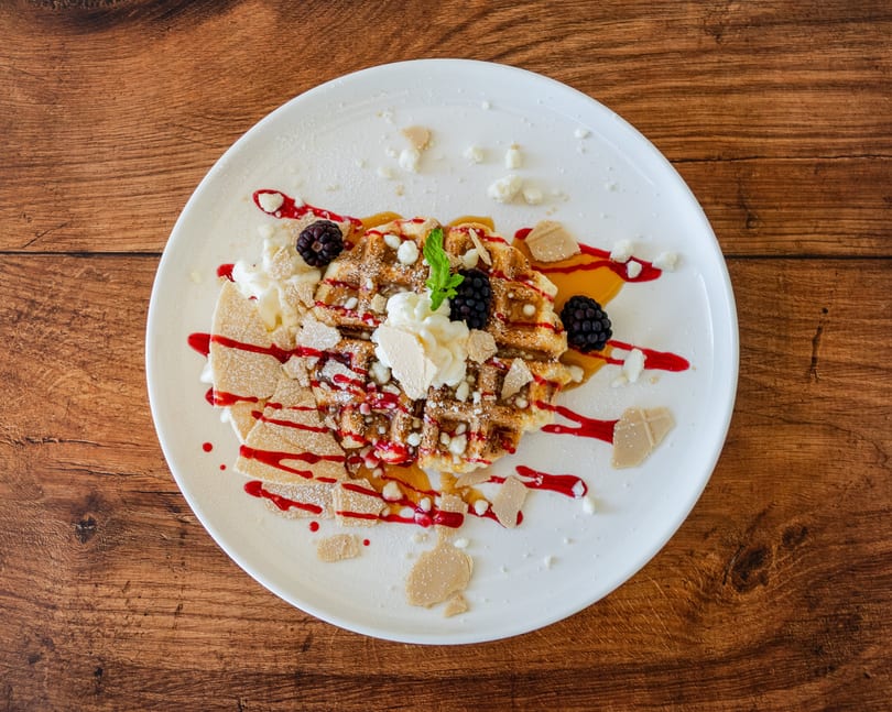 A waffle dessert with whipped cream, blackberries, mint, and berry and caramel drizzles on a white plate.