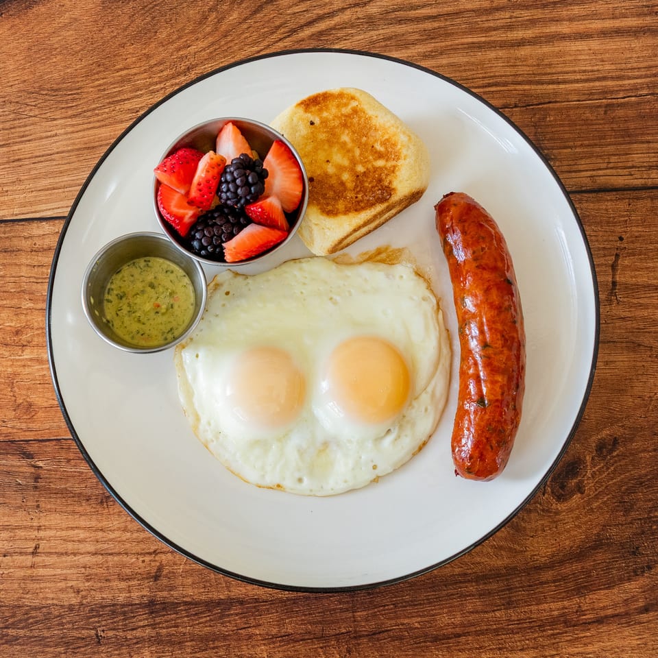 Breakfast plate with two fried eggs, a sausage link, fruit cup, and a toasted biscuit.