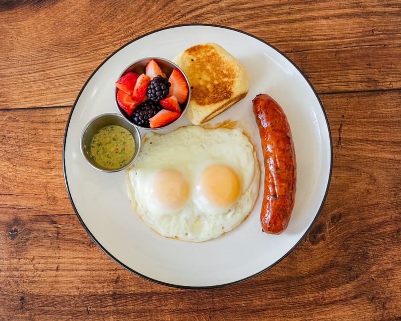 Breakfast plate with two fried eggs, a sausage link, fruit cup, and a toasted biscuit.