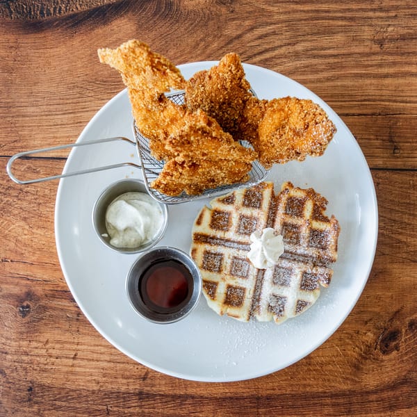 Crispy fried chicken in a metal basket, a waffle with whipped cream, and two dipping sauces.