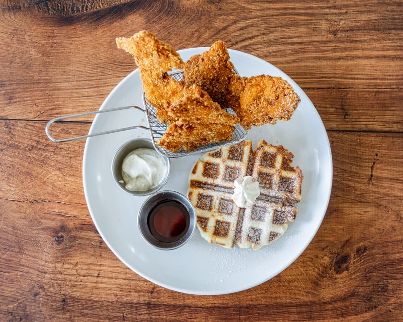 Crispy fried chicken in a metal basket, a waffle with whipped cream, and two dipping sauces.