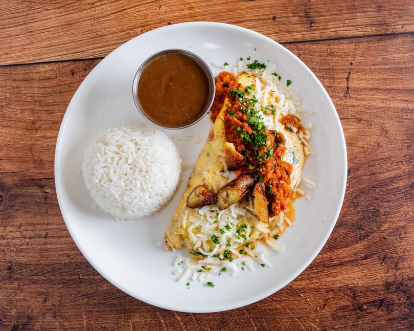 A white plate with an omelet topped with salsa, cheese, plantains, rice, and a side of sauce.
